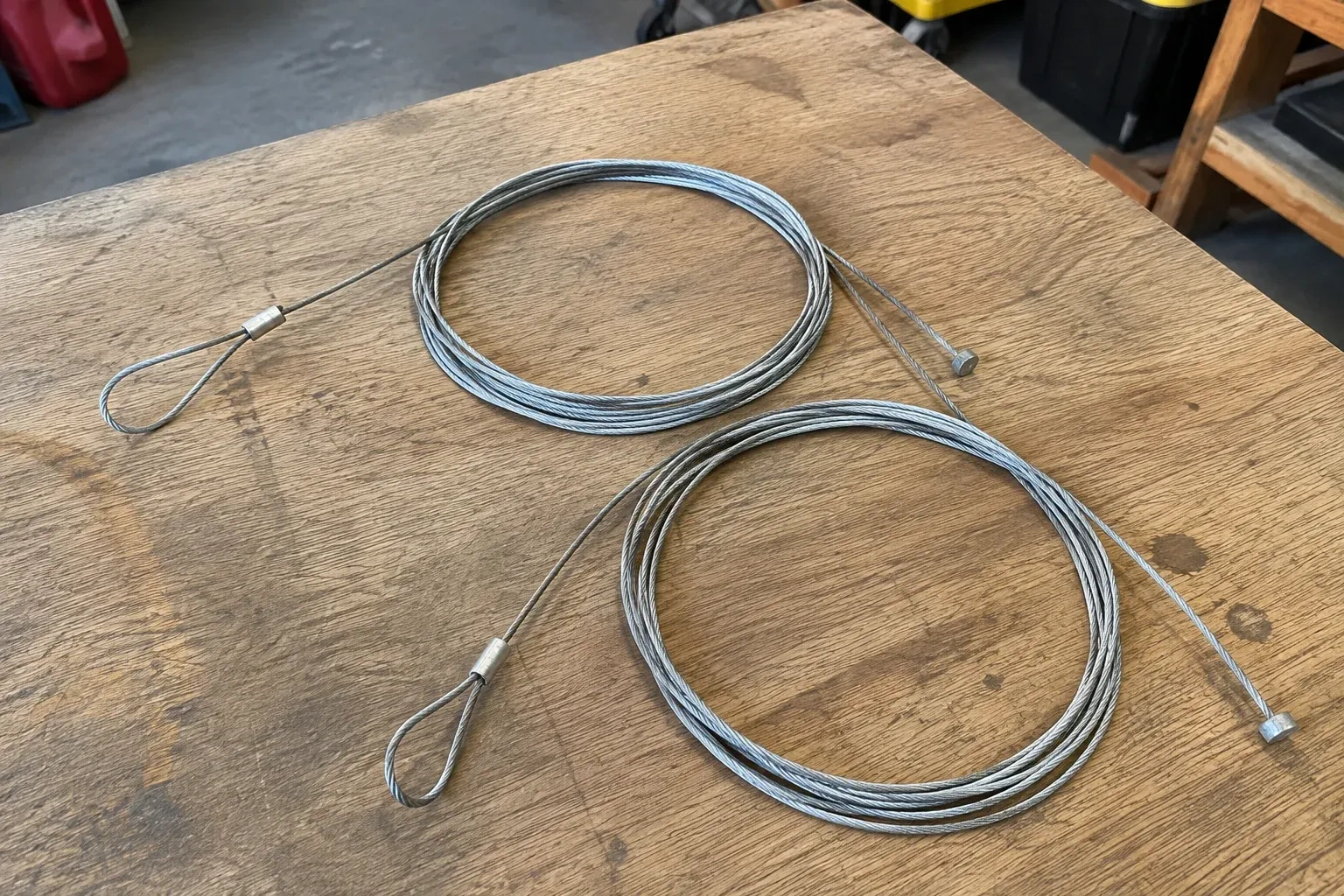 Two coiled steel cables with loop and stop ends on a workbench. — Garage Door Cable Replacement, Burlington