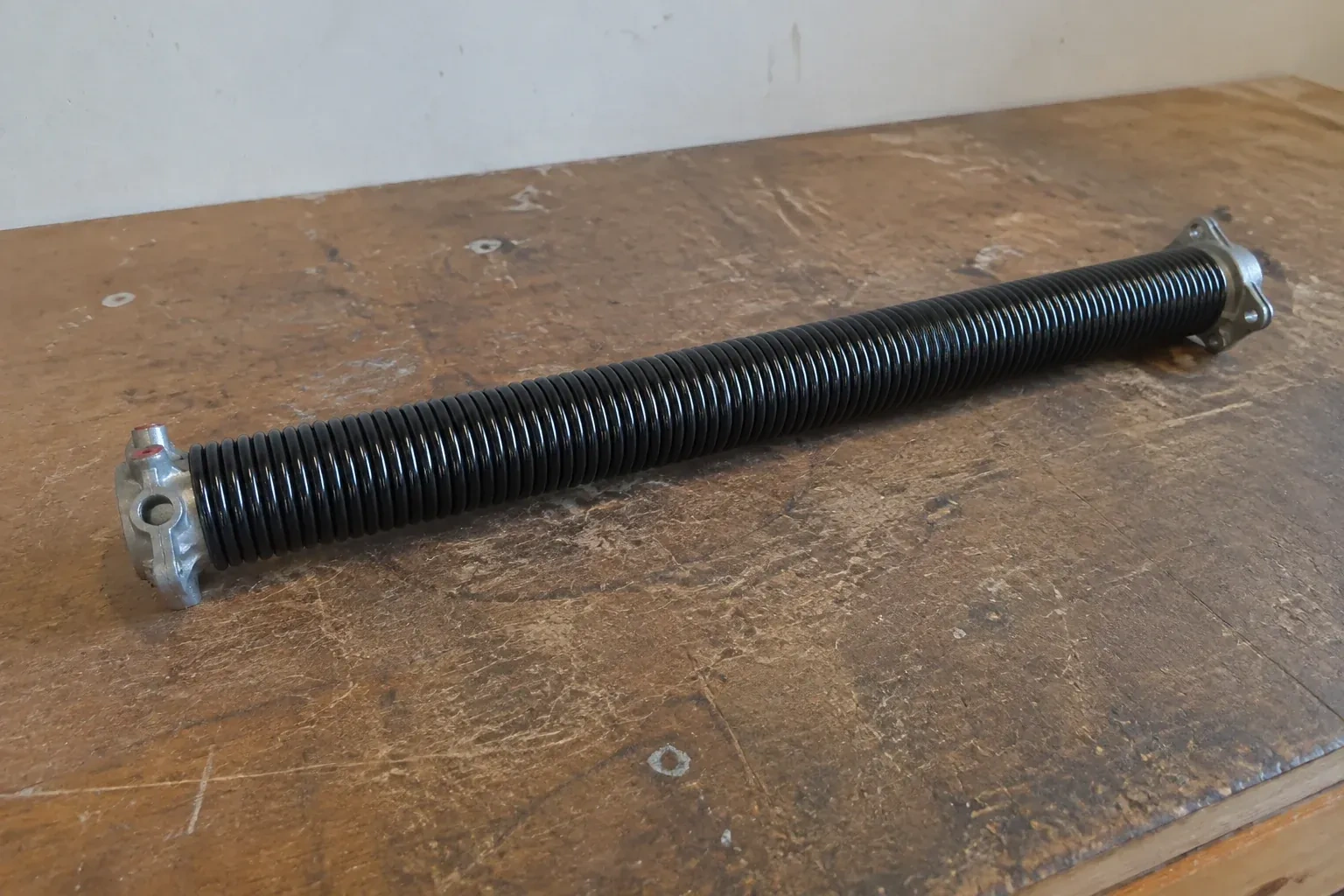 Detached black coil spring with metal end fittings on a workbench. — Torsion Spring Replacement, Burlington