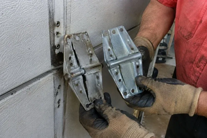 Gloved hands holding two metal garage-door hinges side by side, one discolored with a visible crack and the other bright silver. — Garage Door Hinge Replacement, Burlington